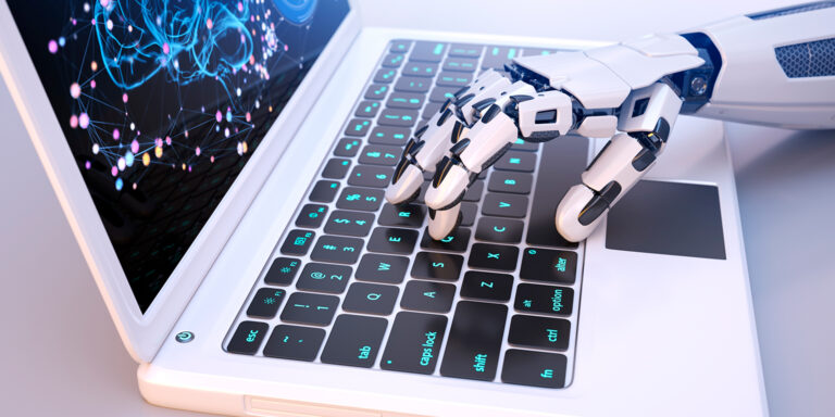Uma mão robótica branca com articulações pretas digitando no teclado de um notebook. Na tela do computador, é exibida a imagem de um cérebro digital azul brilhante formado por redes e pontos luminosos interligados, simbolizando a inteligência artificial