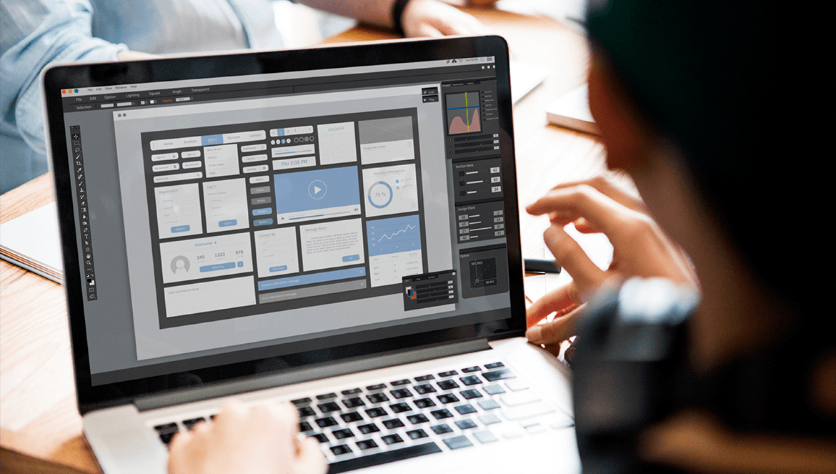 Visão por cima do ombro de uma pessoa trabalhando em um notebook. A tela do computador está em foco e exibe um software de design de interface (UI/UX), onde é possível ver o wireframe de um painel (dashboard) com diversos elementos estruturais, como blocos de layout, gráficos, áreas de perfil e um player de vídeo.