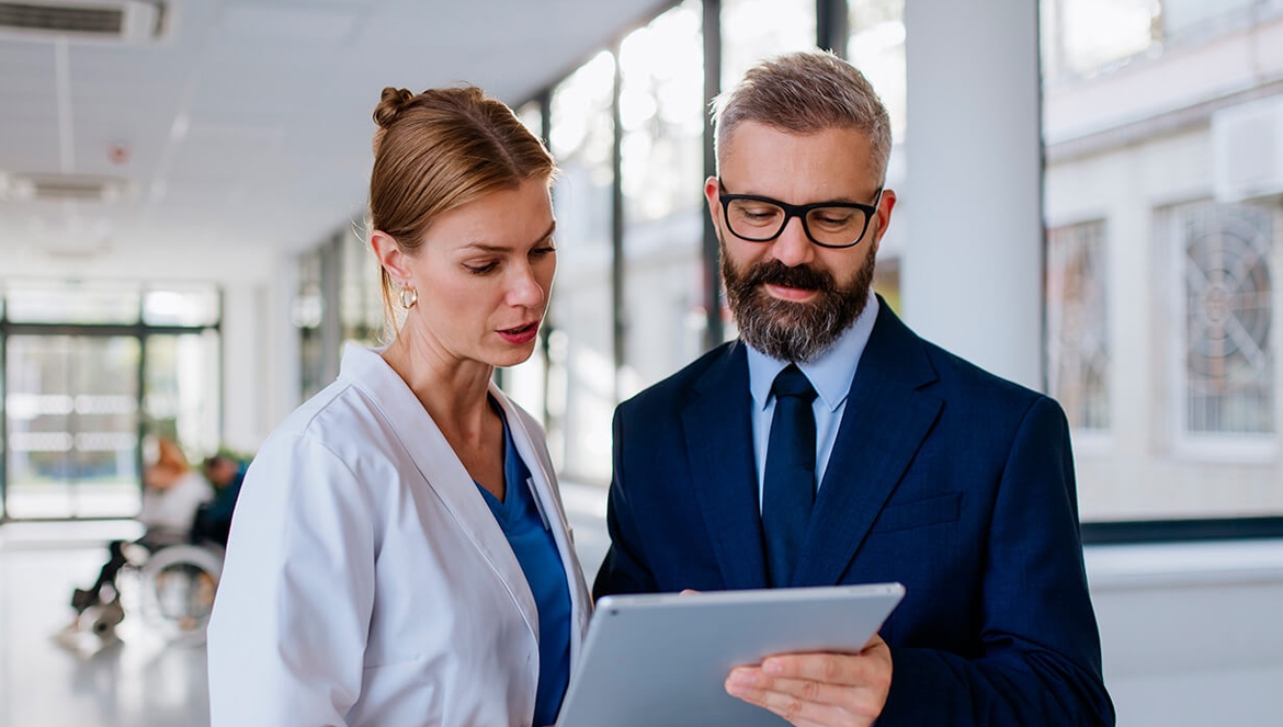 Uma profissional de saúde vestindo jaleco branco e um administrador de terno escuro e óculos olham juntos para a tela de um tablet, discutindo dados em um ambiente clínico iluminado.