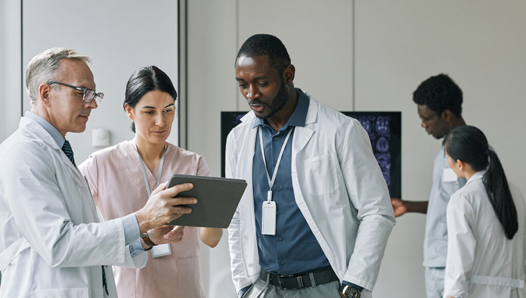 Três profissionais de saúde analisando informações juntos em um tablet em um ambiente clínico moderno.