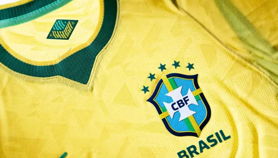 Fotografia em detalhes da camisa amarela da Seleção Brasileira de Futebol. A imagem foca na textura do tecido e destaca o escudo da CBF com cinco estrelas verdes acima e a palavra 'BRASIL' abaixo. A gola em V verde também está visível, com um pequeno emblema na parte interna.